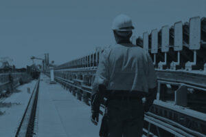 Workman in safety uniform walking beside a coal conveyer belt with coal terminal loaders in the background.