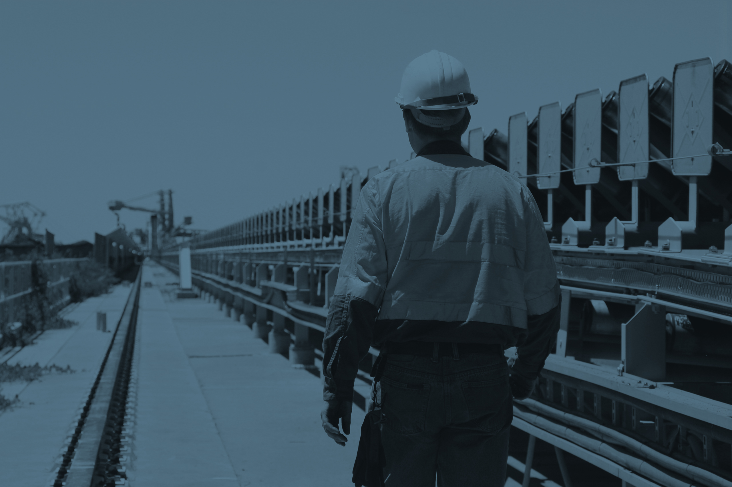 Workman in safety uniform walking beside a coal conveyer belt with coal terminal loaders in the background.